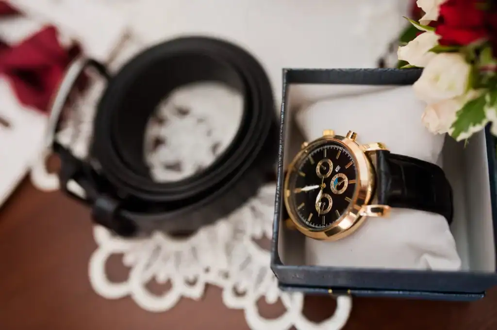 Automatic Watch Winders Explained: From Basic Settings to Pro Tips 2 A luxury wristwatch with a gold case and black leather strap displayed in a gift box, placed next to a black belt and wedding flowers—highlighting the value of storing timepieces in an automatic watch winder to ensure precision and longevity.